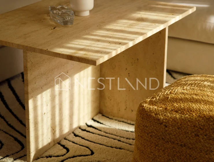 A wooden coffee table with a plant and ashtray, near a textured pouf and rug, in soft lighting.